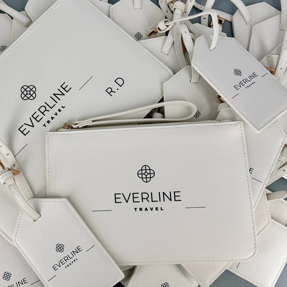 A pile of white Everline Travel branded leather luggage tags, a wristlet pouch, and matching pouches with printed black logos and text, arranged on a gray surface.